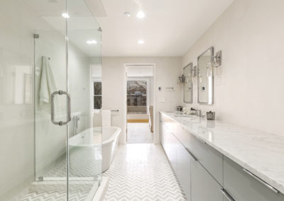 Bright bathroom with chevron floor tiles, glass shower, freestanding tub, and dual vanity with marble top.