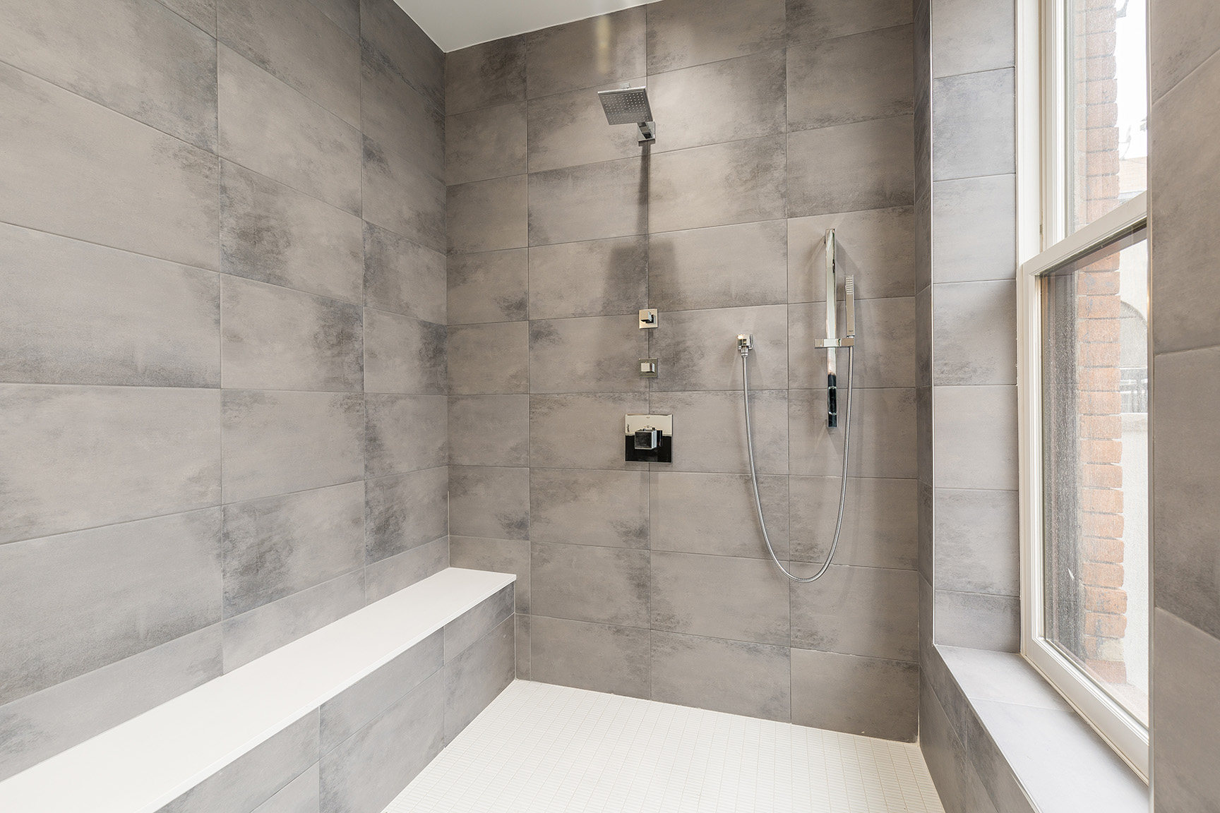 Walk-in shower with large grey tiles and built-in bench, beside a window.