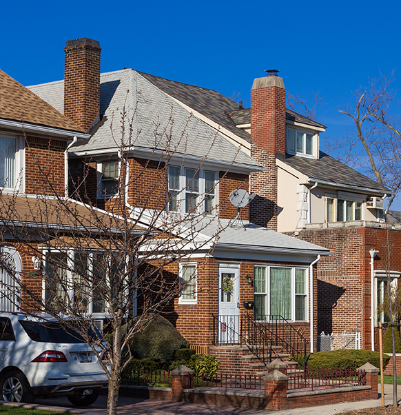 A brick residential home with a steep shingle roof and chimneys under clear skies.