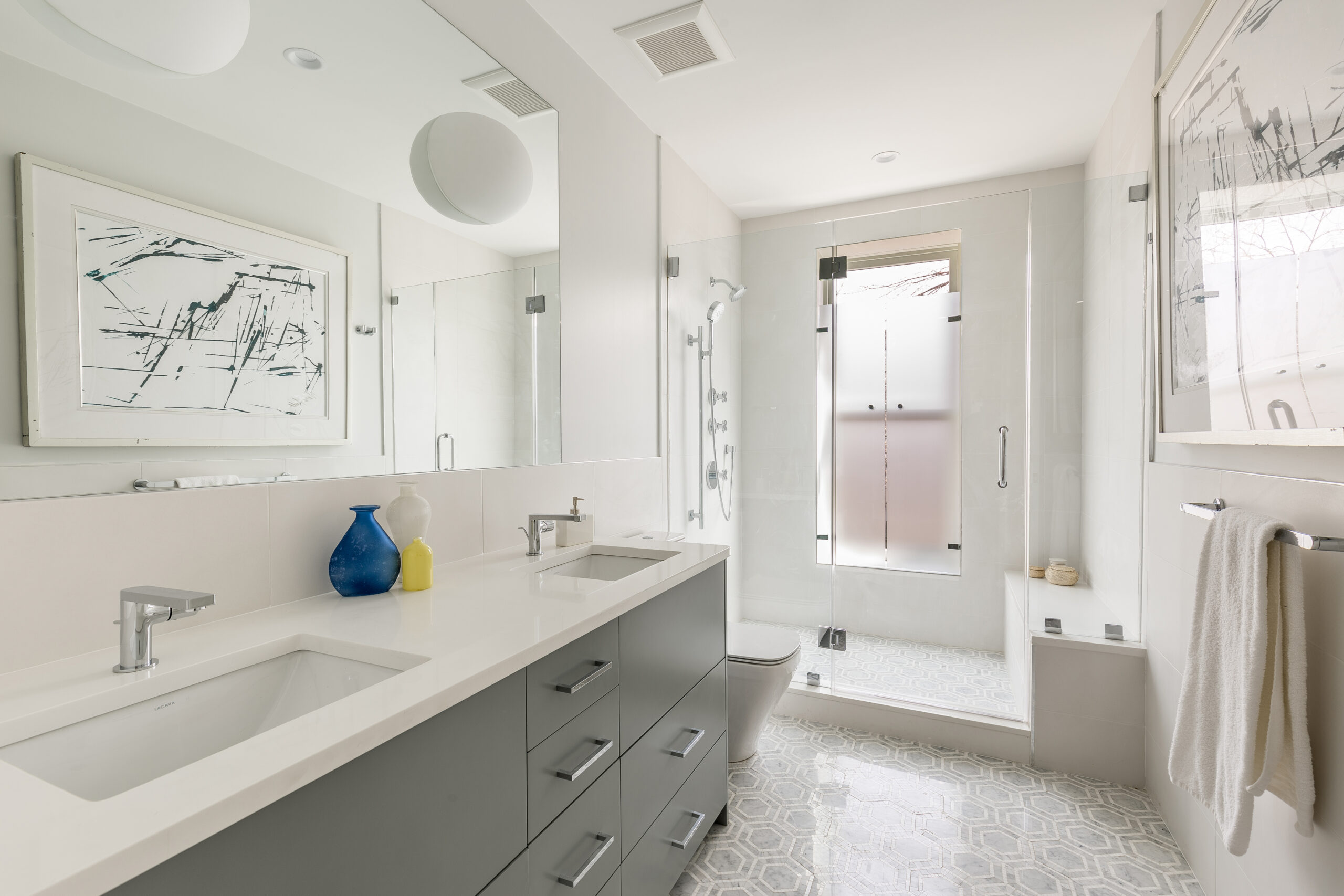 Bright bathroom with dual sinks, glass shower, and abstract art.