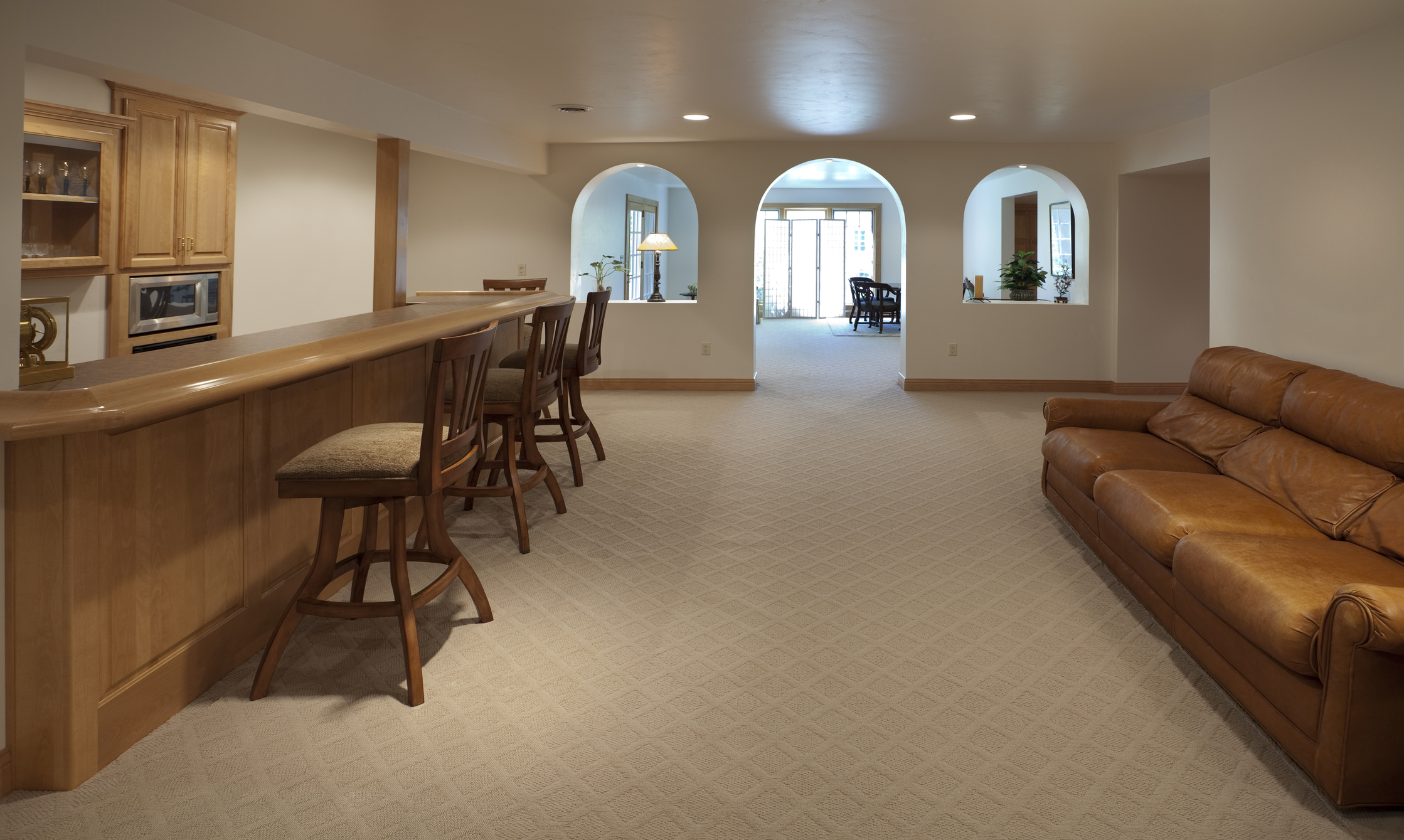 Spacious finished basement with a wooden bar counter, bar stools, and a leather sofa, leading to an archway entrance to another room.