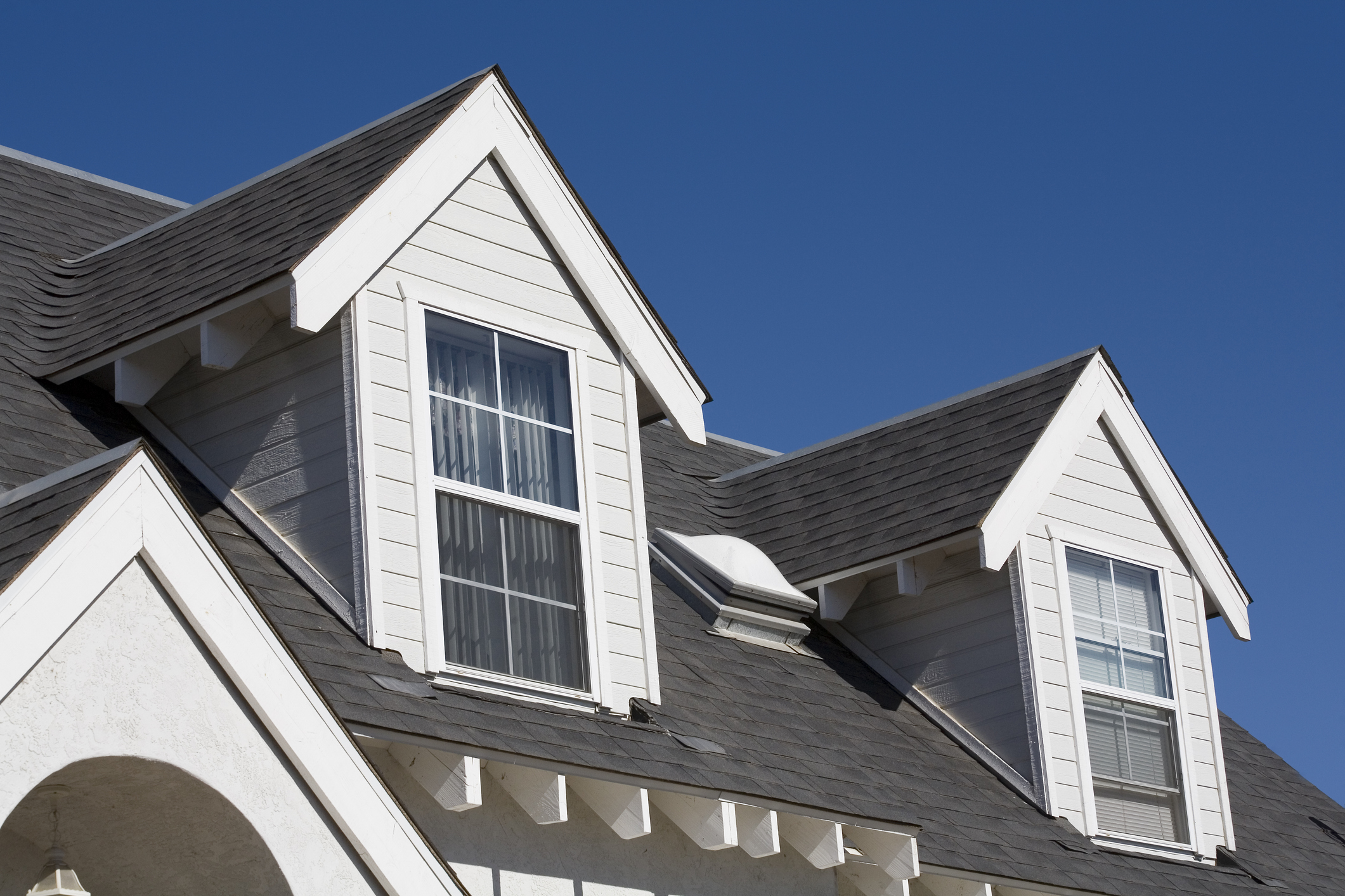 Architectural details of a home's upper level with white dormer windows against a contrasting dark gray shingle roof under a clear blue sky.