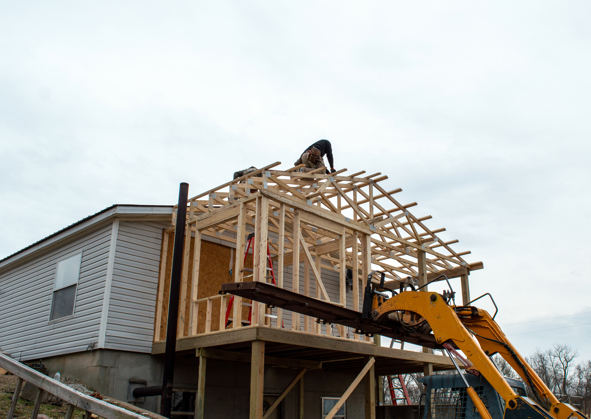 Construction worker on the roof of a home extension in progress, with exposed wooden framing and a telescopic handler nearby.