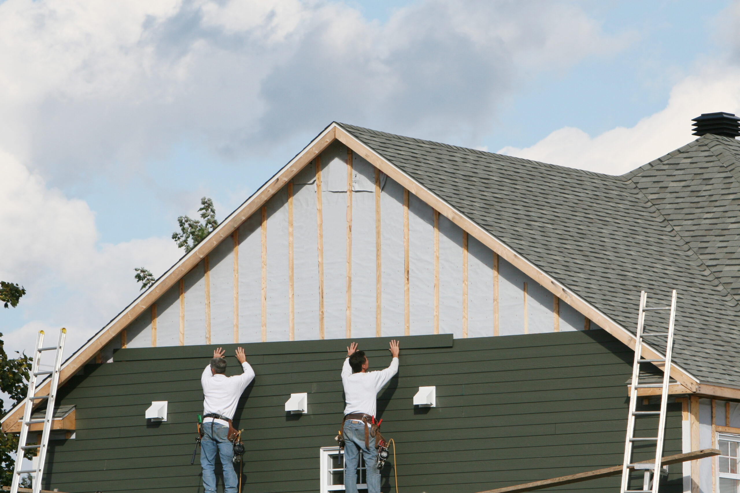 Two workers in white shirts install green siding on the second story of a house, with exposed beams and a gray shingled roof against a cloudy sky.