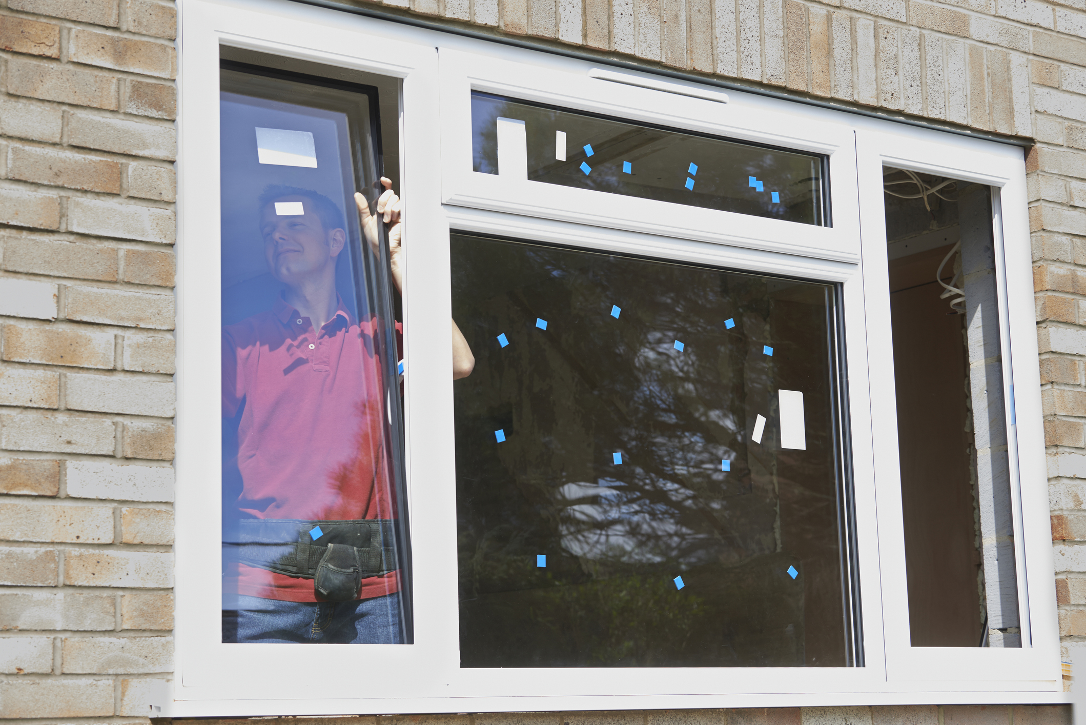 Worker in red shirt installs a new white-framed window with blue tape markers on glass, reflecting trees outside.