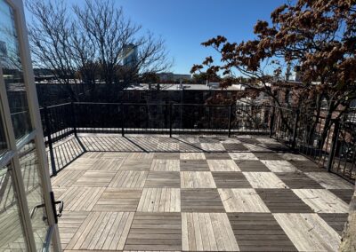 Spacious rooftop terrace with a checkered pattern of wooden deck tiles, surrounded by black metal railings, and offering a clear view of the city skyline under a bright blue sky.