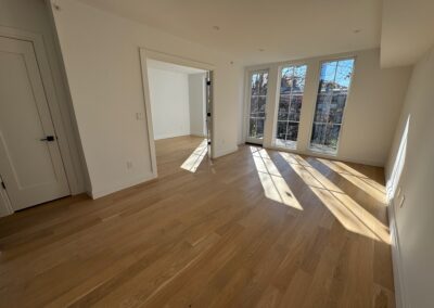 Spacious and sunlit room with light wood flooring, large floor-to-ceiling windows, and an open doorway leading into a bright adjacent room, highlighting a modern residential renovation.