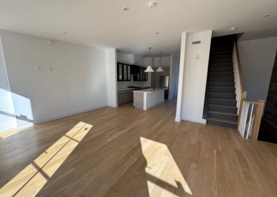 Modern open-concept living space with light hardwood floors, a sleek kitchen featuring a central island and pendant lighting, and a staircase with white spindles leading to the upper level. Natural light fills the room through large windows.
