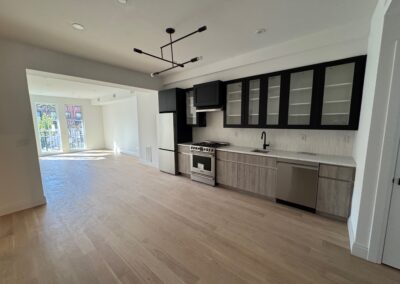 Modern kitchen with light wood lower cabinets, black-framed glass upper cabinets, and stainless steel appliances, including a refrigerator, oven, and dishwasher. The space features a minimalist black faucet and a linear light fixture overhead. The open layout extends into a bright living area with large windows and natural light.